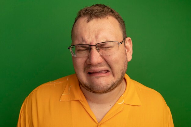Расстроенный мужчина в очках и оранжевой рубашке, плачущий с безнадежным выражением лица, стоит над зеленой стеной