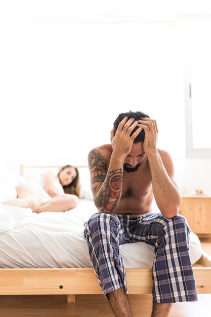 Upset man sitting on bed in front of his wife