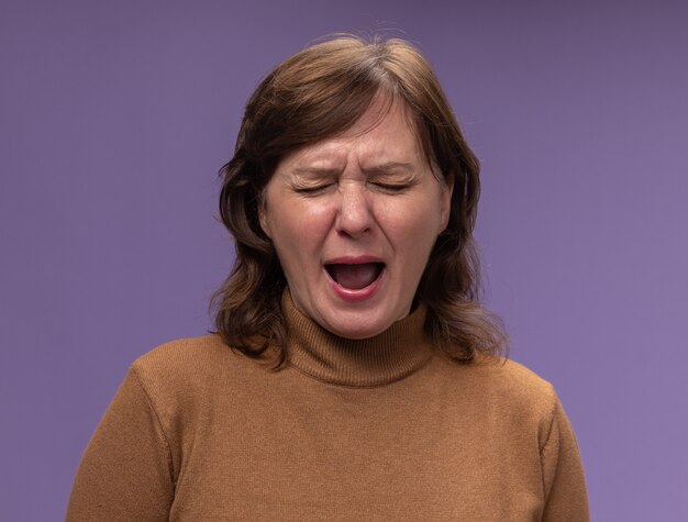 Upset middle aged woman in brown turtleneck crying hard with closed eyes standing over purple wall