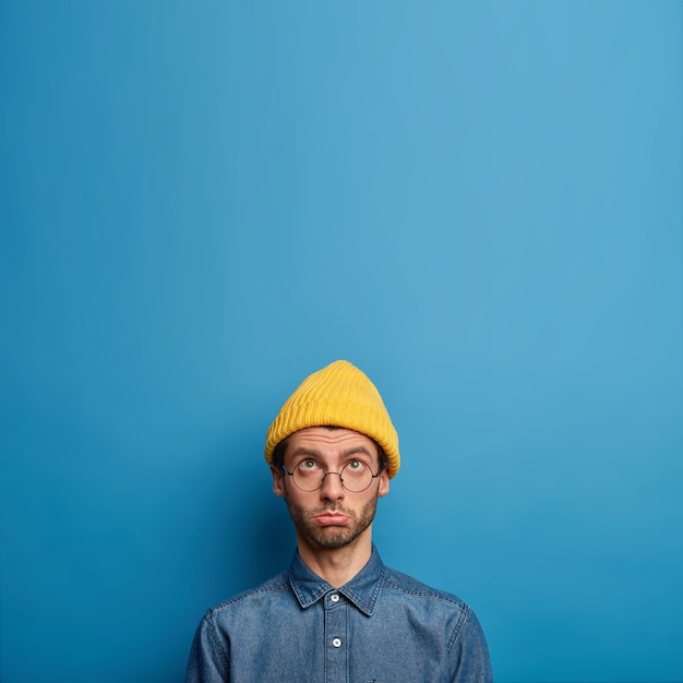 Upset offended man focused above, complains of hard life, wears yellow hat and denim shirt, looks up with sad face