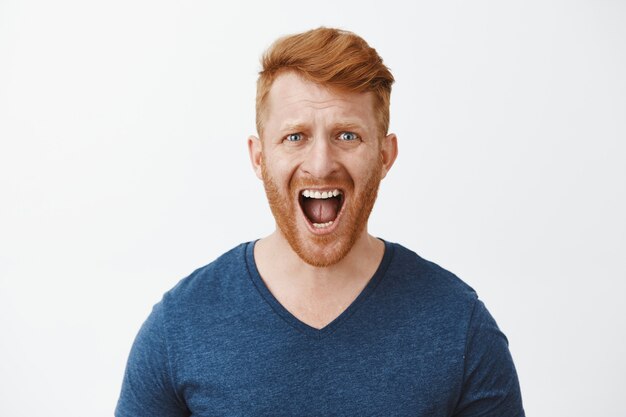 Upset outraged and unhappy redhead man with bristle, shouting, frowning making displeased expression, feeling pain in soul while having argumbent over grey wall