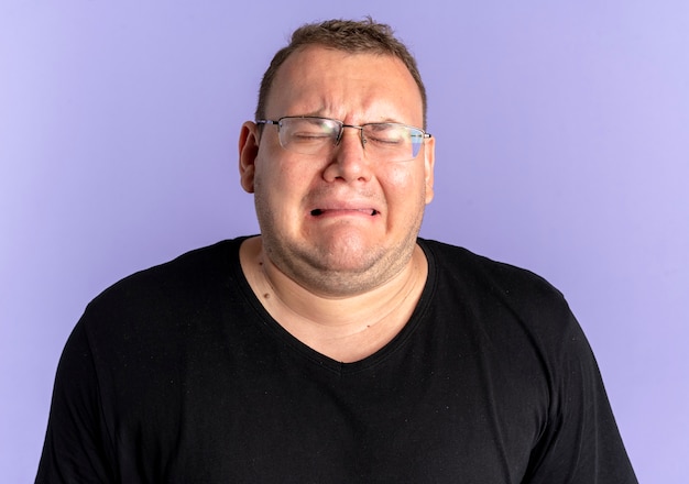 Upset overweight man in glasses wearing black t-shirt with unhappy face going to cry standing over blue wall