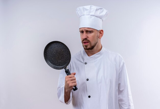 Upset professional male chef cook in white uniform and cook hat holding a pan with sad expression on face standing over white background