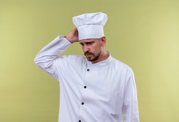 Cuoco chef maschio professionista sconvolto in uniforme bianca e cappello da cuoco che sembra stanco e oberato di lavoro toccando la sua testa in piedi su sfondo gree