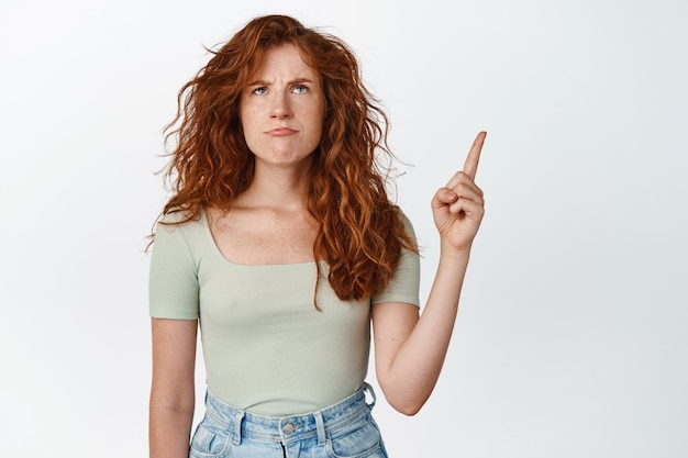 Upset redhead woman grimacing displeased pointing and looking up with angry face expression standing over white background