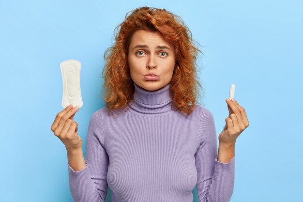 Upset redhead woman holds hygienic sanitary pad and tampon, chooses good protection during red days, has gloomy face expression, wears casual jumper, isolated on blue wall. Feminity concept