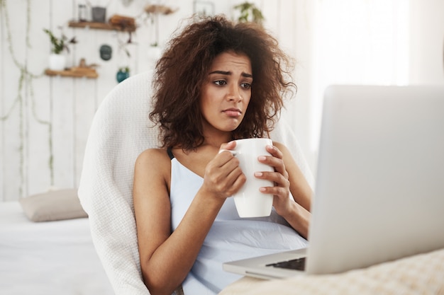 Upset sad beautiful african woman looking at laptop holding cup sitting in chair at home spending her weekend alone.