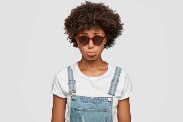 Upset sad young African American female purses lower lip, feels abused, wears fashionable round sunglasses and denim overalls, poses against white wall. People, emotions and style concept