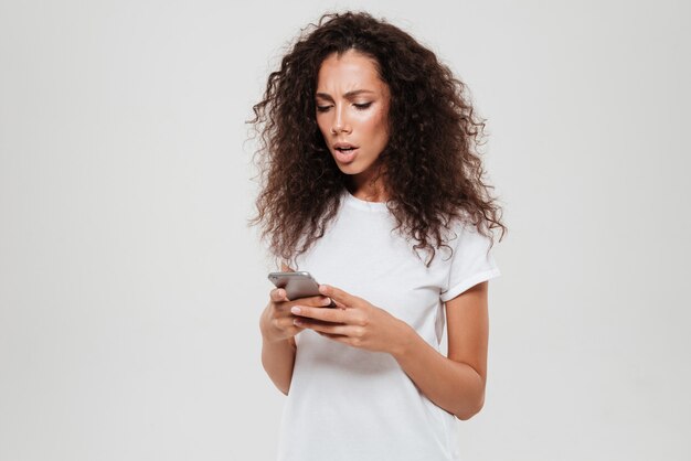 Upset serious curly woman writing message on smartphone