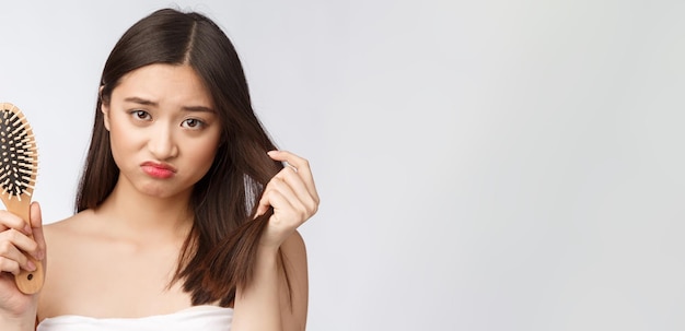 Upset stressed young Asian woman holding damaged dry hair on hands over white isolated background