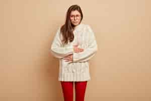 Free photo upset sullen european female touches belly from pain, feels unwell, discomfort after eating spoiled product, wears spectacles and warm clothes, stands over brown wall. stomachache concept