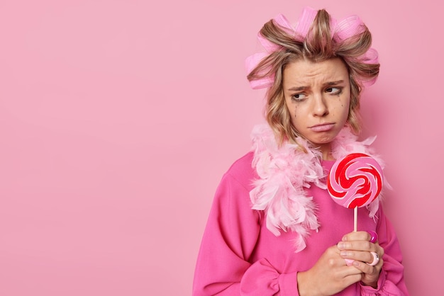 Foto gratuita la donna premurosa sconvolta ha perso il trucco tiene il lecca-lecca rotondo sul bastone mantiene la dieta non può mangiare dolci applica i bigodini per fare il taglio di capelli indossa pose di abiti festivi su sfondo rosa