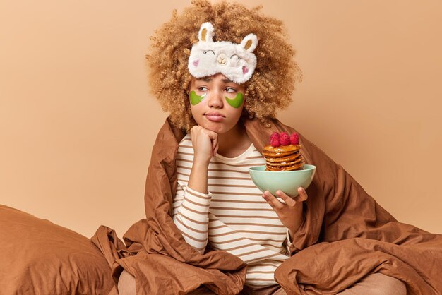 곱슬머리를 한 화난 사려깊은 여성은 담요 아래 침대에 누워 있고 맛있는 식욕을 돋우는 팬케이크 그릇을 들고 눈 아래 녹색 하이드로겔 패치를 적용합니다.