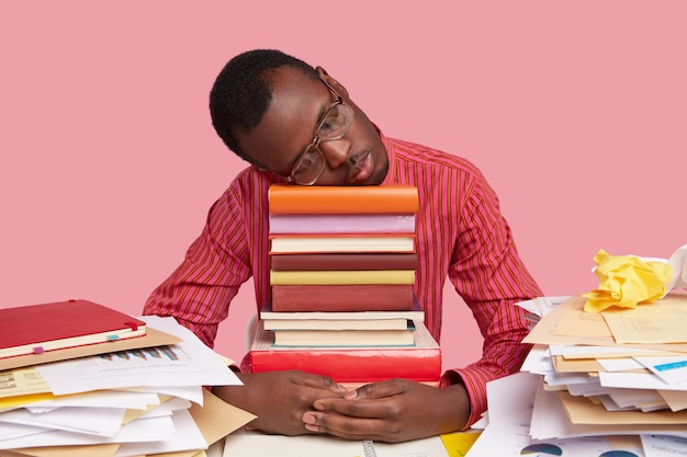 Free photo upset tired black man takes nap on pile of books, sleeps after studying all night, prepared for exams