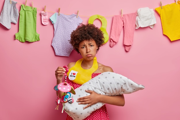 Upset tired mother poses with newborn baby, holds mobile toy, bib on neck, busy nursing infant, needs help of husband, plays and feeds small recently born child. Postnatal depression, mood disorder