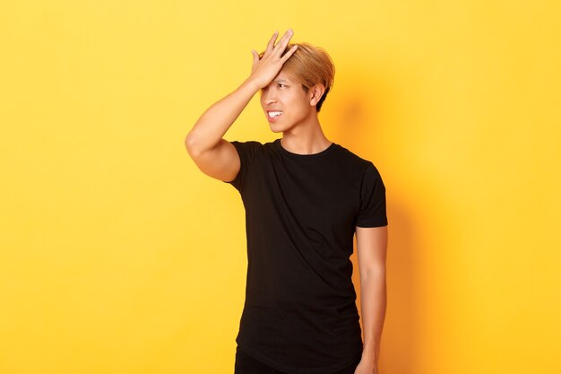 Upset and troubled asian guy snap forehead forgetful, standing over yellow wall