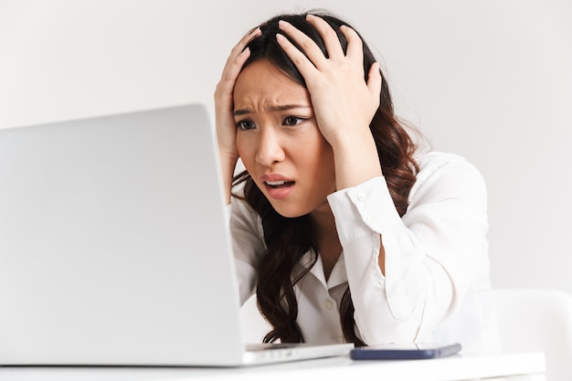 ノート​パソコン​を​見て​、​手​で​頭​を​つかんで​オフィス服​を​着ている​動揺​の​不幸な​アジア​ビジネス​女性
