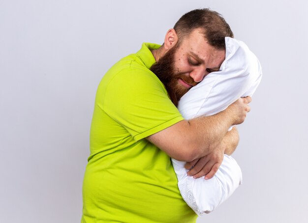 Upset unhealthy bearded man in yellow polo shirt feeling sick holding pillow wants to sleep standing over white wall