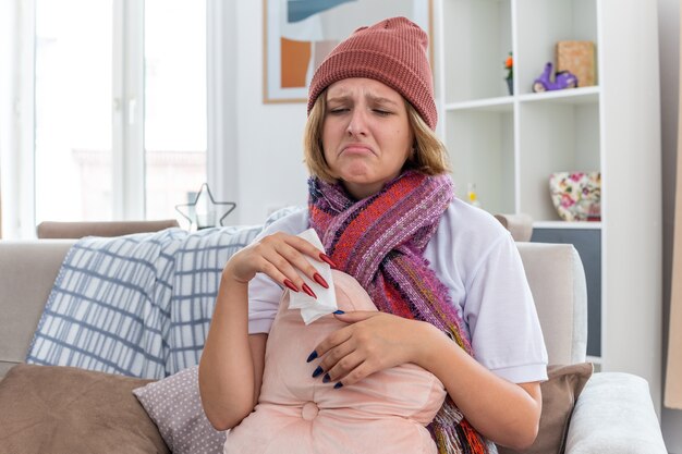 Расстроенная нездоровая молодая женщина в теплой шляпе с шарфом, которая выглядит нездоровой и больной, держит ткань, страдающую от простуды и гриппа, и сморкается, сидя на диване в светлой гостиной