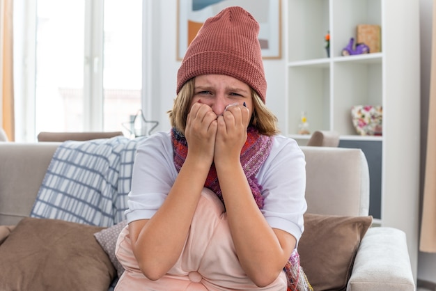 Расстроенная нездоровая молодая женщина в теплой шляпе с шарфом, которая выглядит нездоровой и больной, держит ткань, страдающую от простуды и гриппа, и беспокоит сморкание, сидя на диване в светлой гостиной