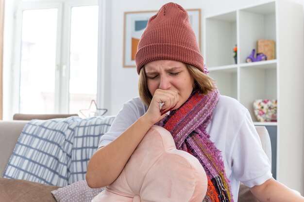 Upset unhealthy young woman in warm hat with scarf looking unwell and sick holding tissue suffering from cold and flu and coughing sitting on couch in light living room