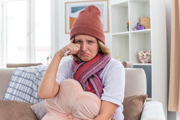 Расстроенная нездоровая молодая женщина в теплой шляпе с шарфом, которая выглядит нездоровой и больной, держит ткань, страдающую от простуды и гриппа, плачет, протирая глаза, сидя на диване в светлой гостиной