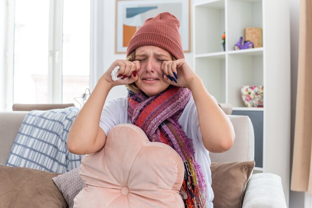 Расстроенная нездоровая молодая женщина в теплой шляпе с шарфом, которая выглядит нездоровой и больной, держит ткань, страдающую от простуды и гриппа, плачет, протирая глаза, сидя на диване в светлой гостиной
