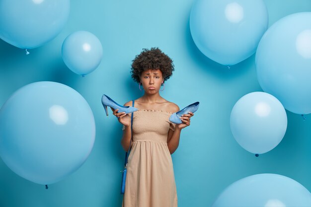 Upset woman got calluses while wearing high heel shoes, wears cocktail dress, has bad mood, tired after party, isolated on blue wall decorated with inflated balloons. Women clothes collection
