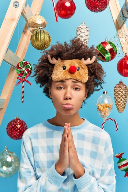 Upset woman keeps palms pressed together pleads for favor wears pajama and sleepmask needs help in decorating house for winter holidays poses around new year toys isolated over blue background