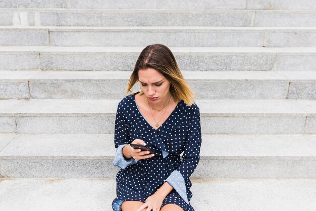 Upset woman sitting on staircase looking at smartphone