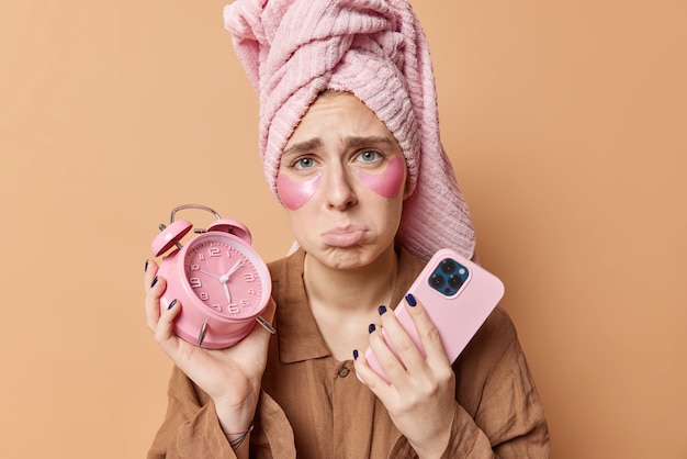 Upset woman wakes up early in morning holds alarm clock and smartphone undergoes beauty procedures daily routines wears pajama wrapped towel on head isolated over brown background. Awakening