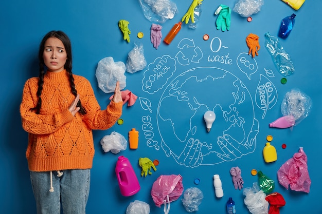 Upset worried young girl in casual clothes shows no gesture against plastic waste, gestures against blue background with drawn globe