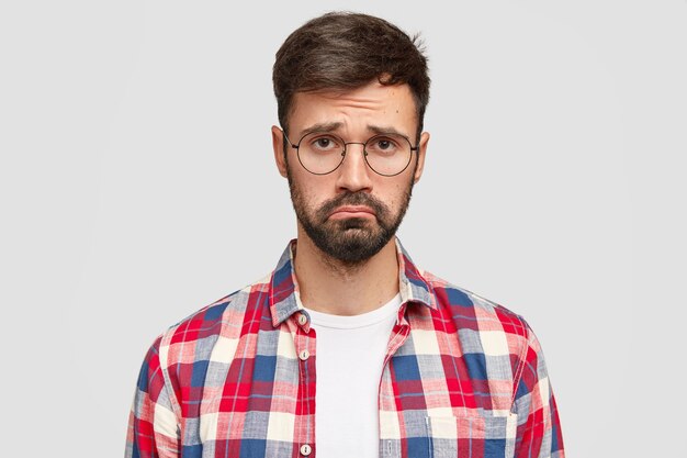 Upset young bearded man with gloomy expression, being offended by someone, raises eyebrows