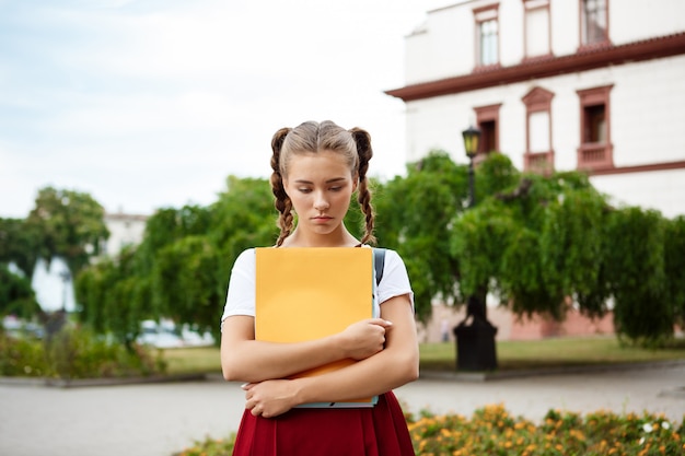 Upset young beautiful female student looking down holding folders outdoors