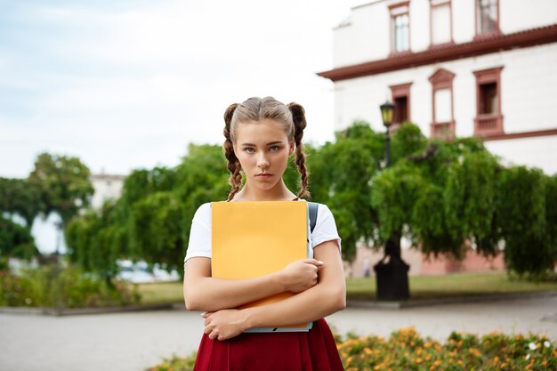 Upset young beautiful female student looking and holding folders outdoors