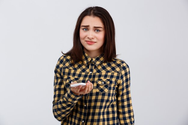 Upset young beautiful girl dressed in plaid shirt holding phone, over white wall.