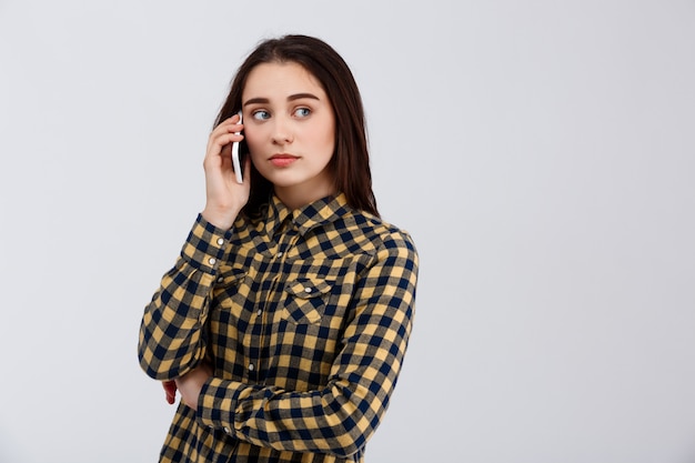 Upset young beautiful girl dressed in plaid shirt speaking on phone, looking at side over white wall.