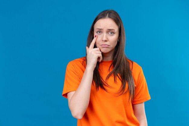 Upset young beautiful girl wearing orange t-shirt pointing with finger to her eye looking at camera with sad expression face standing over isolated blue background