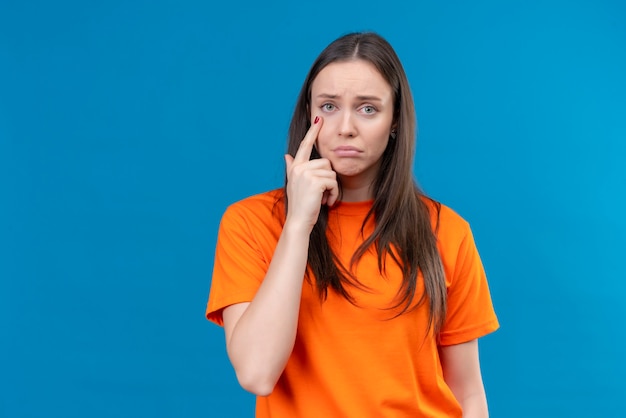 Расстроенная молодая красивая девушка в оранжевой футболке, указывающая пальцем в глаз, смотрит в камеру с грустным выражением лица, стоящим на изолированном синем фоне