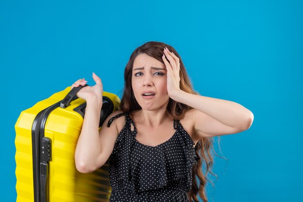 Upset young beautiful traveler girl in dress in polka dot holding suitcase looking at camera with sad expression on face standing over blue background