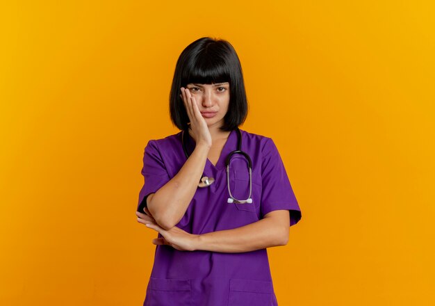 Upset young brunette female doctor in uniform with stethoscope puts hand on face