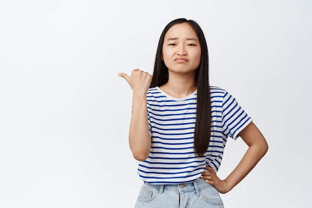 Upset young brunette woman pointing finger left and sulking with regret, showing smth upsetting, dislike, standing on white.