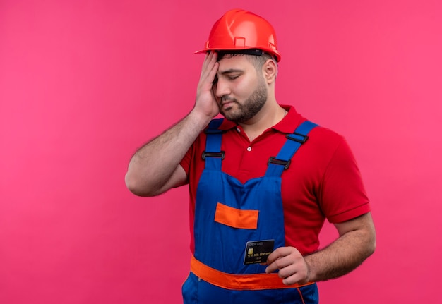 Upset young builder man in construction uniform and safety helmet holding credit card looking confused 