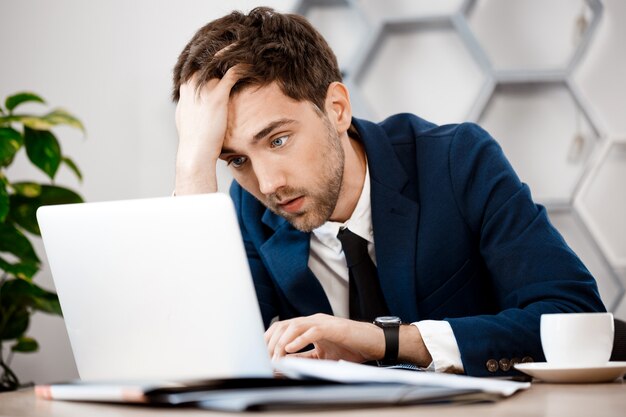 ノートパソコン、オフィスの背景に座っている青年実業家を混乱させます。