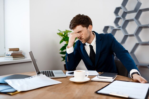 Upset young businessman sitting at workplace, office background.