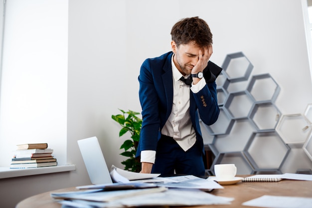 Upset young businessman standing at workplace, office background.