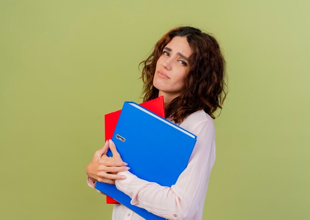 Upset young caucasian girl holds file folders looking at camera isolated on green background with copy space