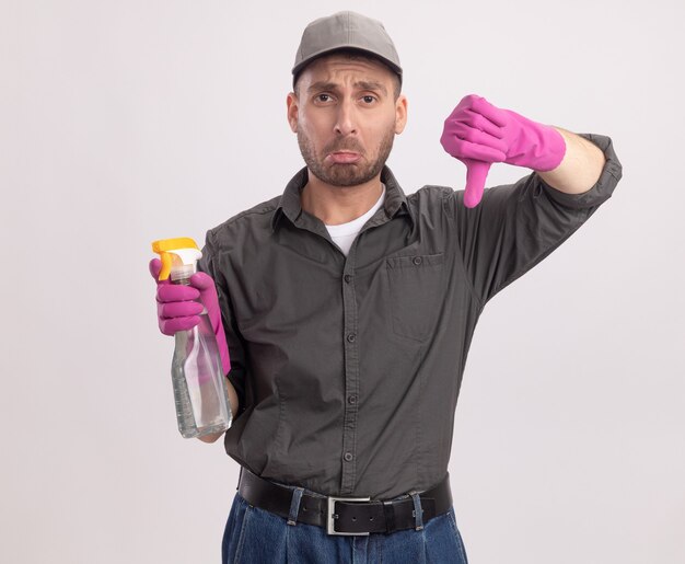 Upset young cleaning man wearing casual clothes and cap in rubber gloves holding spray bottle looking  with sad expression showing thumbs down standing over white wall