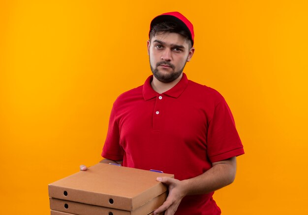 Upset young delivery man in red uniform and cap holding stack of pizza boxes looking at camera with sad expression on face