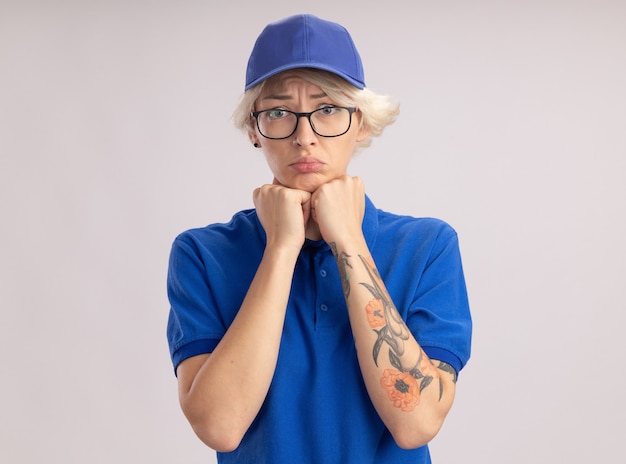 Sconvolto giovane donna di consegna in uniforme blu e cappello guardando con espressione triste sul viso in piedi sopra il muro bianco